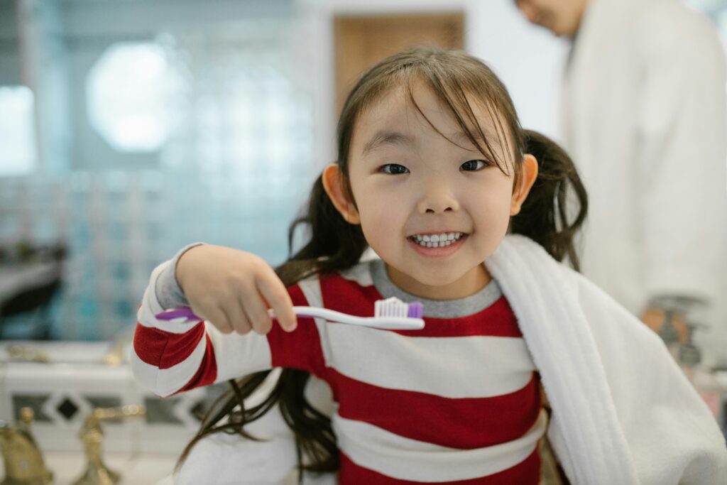 A smiling young girl in a bathroom holding a toothbrush, promoting dental hygiene.